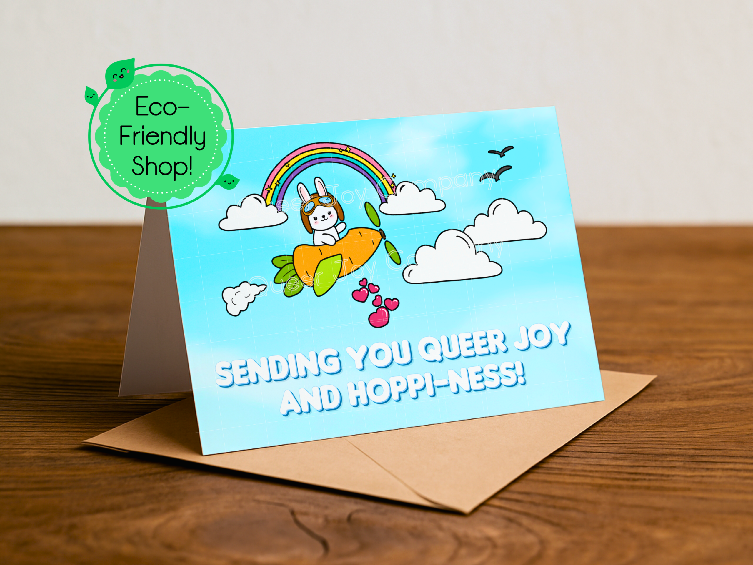 A cute kawaii bunny in a carrot airplane flies beneath a rainbow and clouds delivering hearts with a title saying Sending You Queer Joy and Hoppi-ness